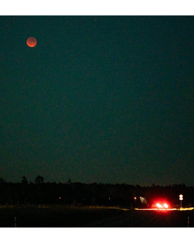 An Image of a "Blood Moon" with a car driving through a countryside. The full Moon eclipse.