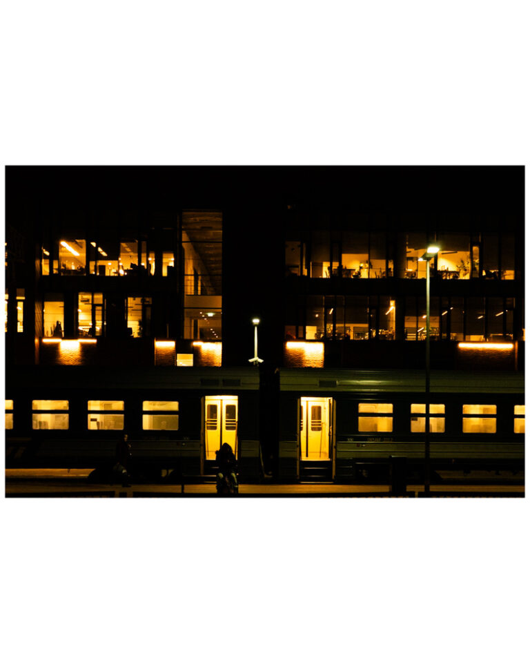 A photo of a train station. Late evening. The trains doors are open, yellow lights pours outside. A person sits in front of the door on a bench. Only a silhouette of this person is visible.